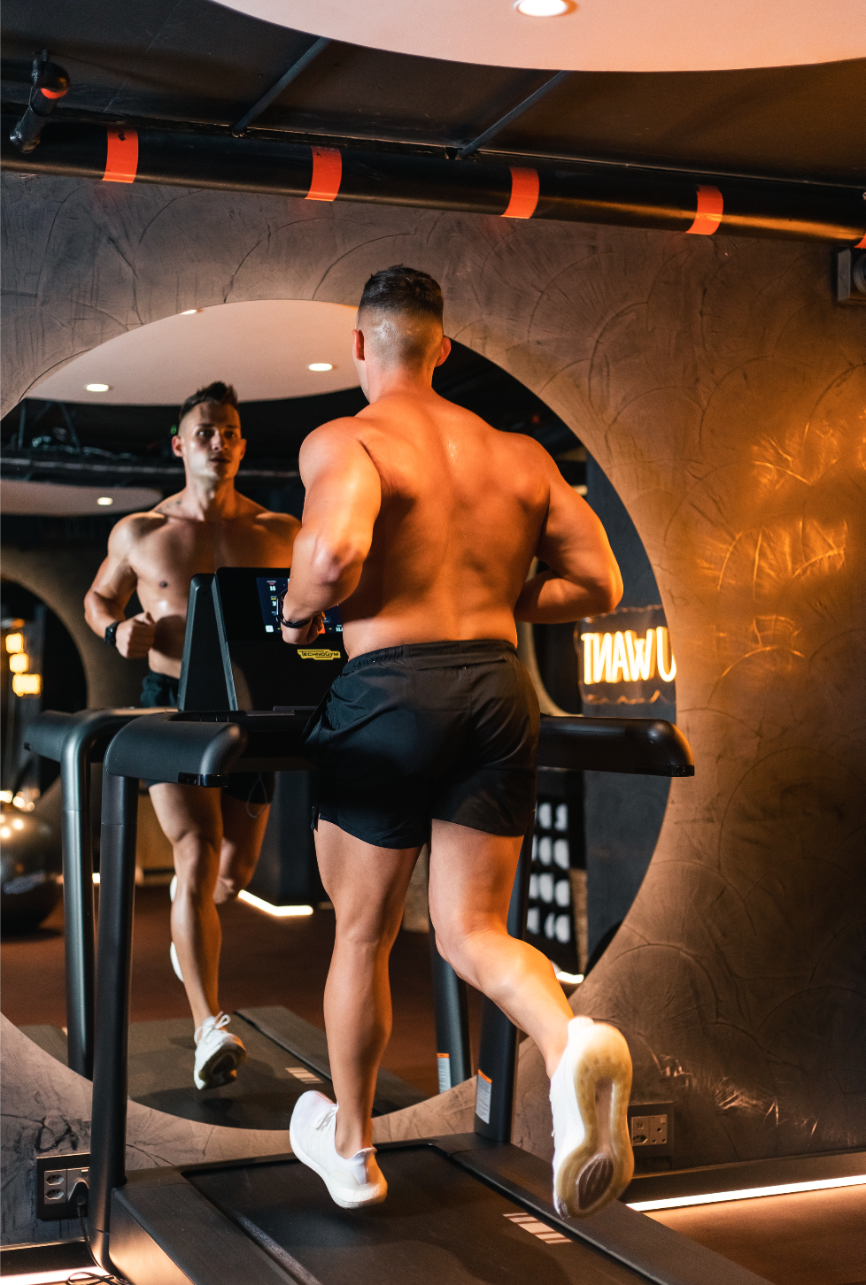 A shirtless man running on a treadmill. The room is lit with warm yellow light. There is a large circular mirror in front of him that he is looking into.