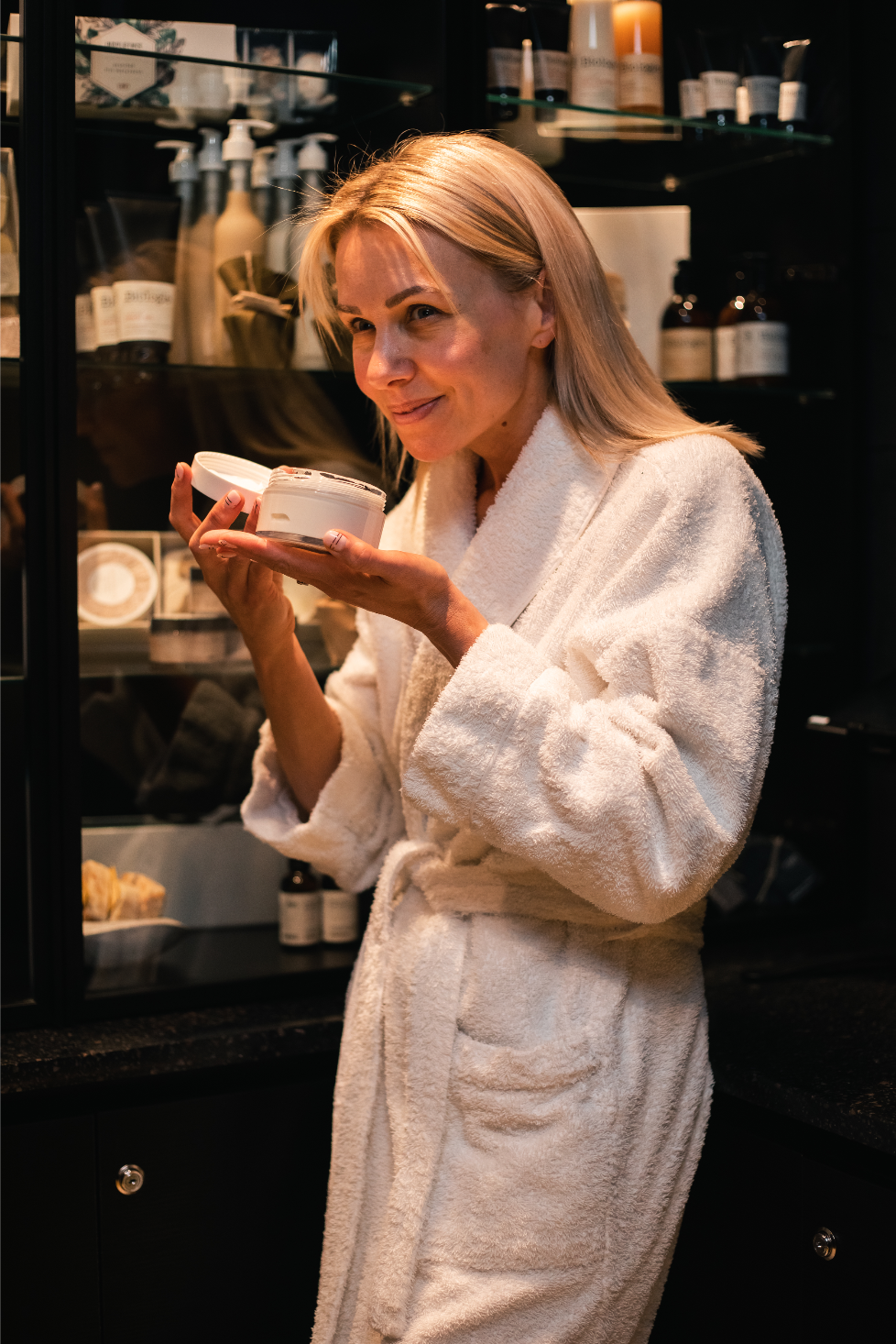 A woman in a white bathrobe holding and smelling a jar of cream, with a glass cabinet displaying skincare products behind her